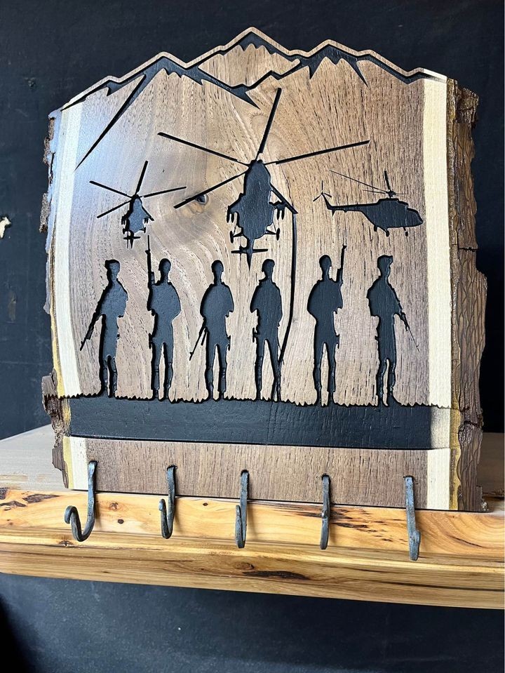 Wooden plaque with silhouettes of soldiers and helicopters against a mountain background, mounted above metal hooks.